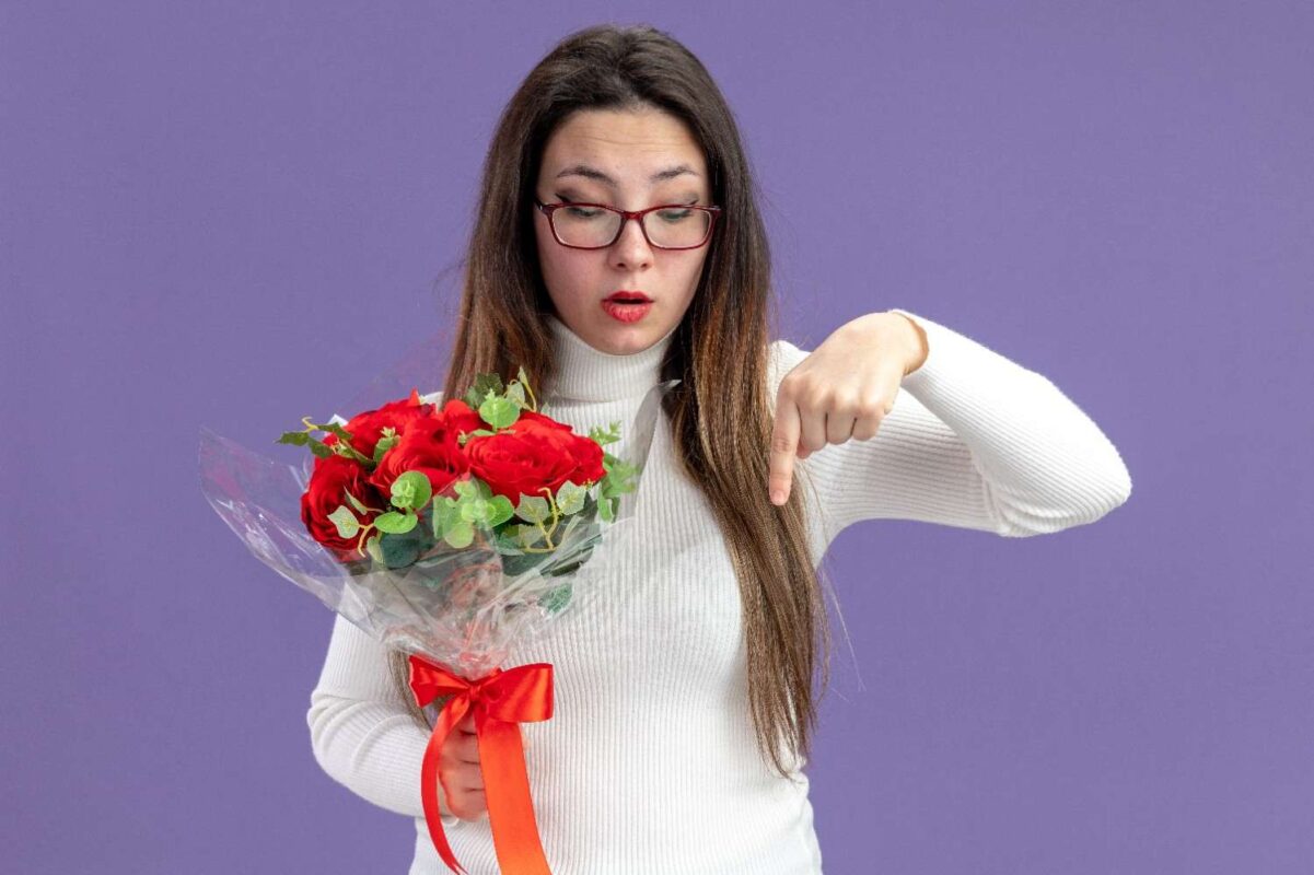 A young beautiful woman wearing casual clothes holding bouquet red roses looking down pointing with index finger down to indicate mistakes in flower bouquet.